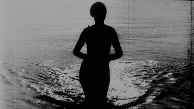 Black and white archive footage. The silhouette of a woman rises from the ocean.