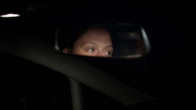 The darkened dashboard of a car. The eyes of a person sitting in the back seat are reflected in the rearview mirror.