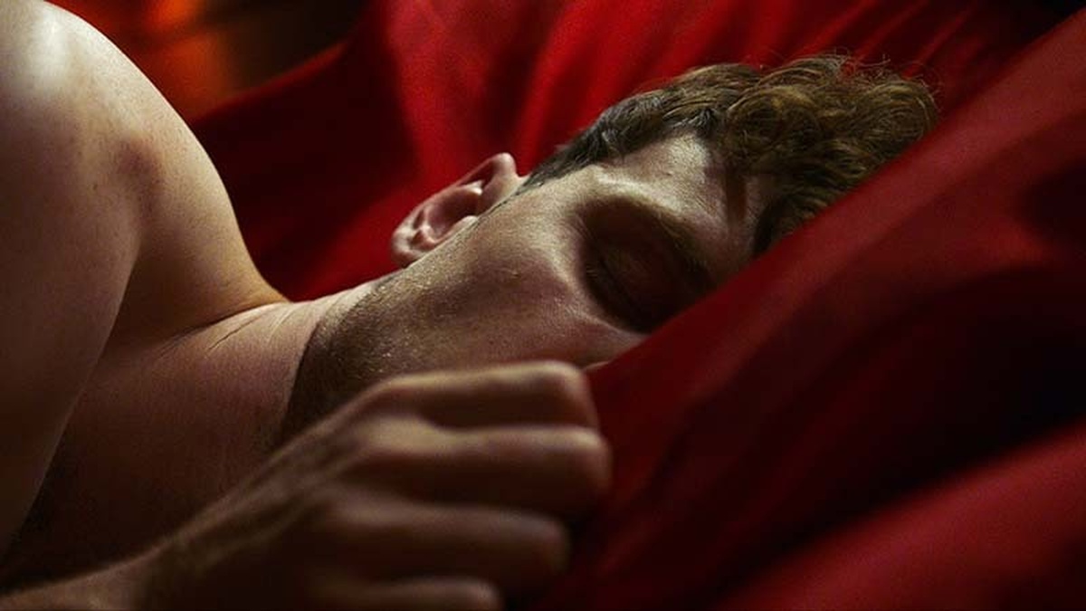 A man sleeps on red bedding. His hand rests on the pillow and partially obscures his face.