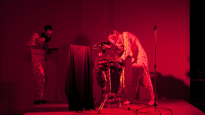 There are two people on a red tinged stage both wearing pyjama-like costumes, holding a glove puppet and a theremin.