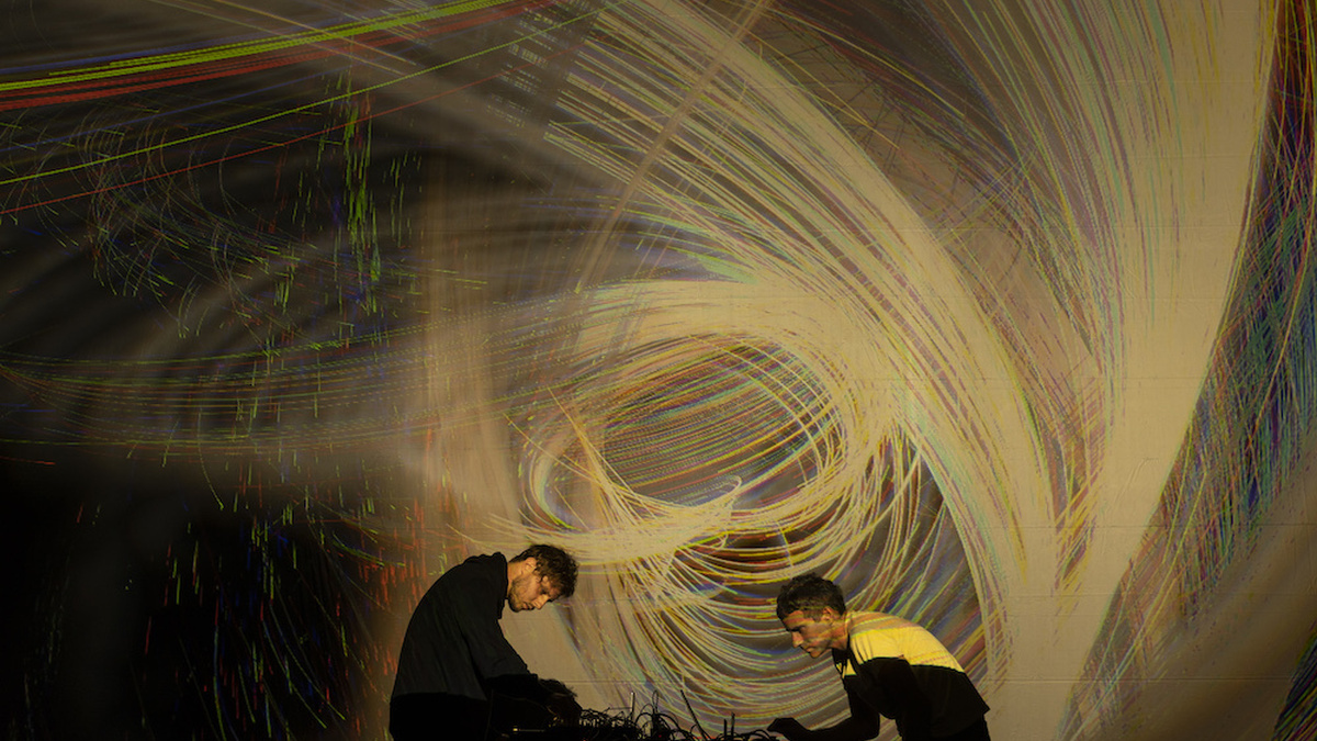 Artists Maotik and Maarten Vos perform in front of a large screen projecting a swirling storm-like typhoon graphic.