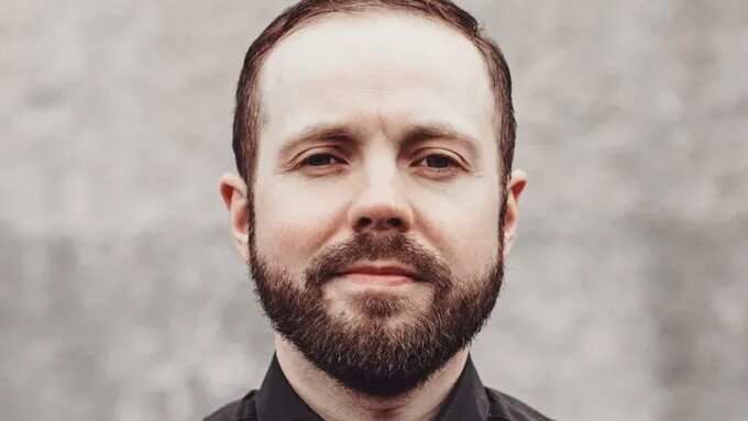 A person with brown hair and a beard wears a black shirt and holds a guitar.