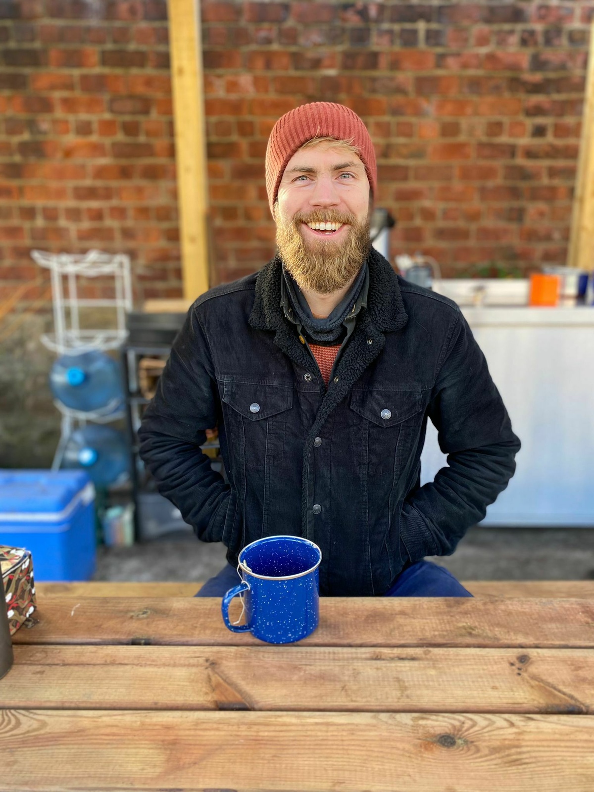 a bearded smiling person in a red beanie hat, sitting at a sunny bench outdoors with a big blue mug of tea