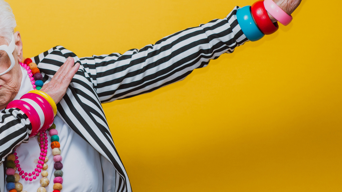 An older person dressed in a striped jacket and jewellery performing a dance move against a yellow background.