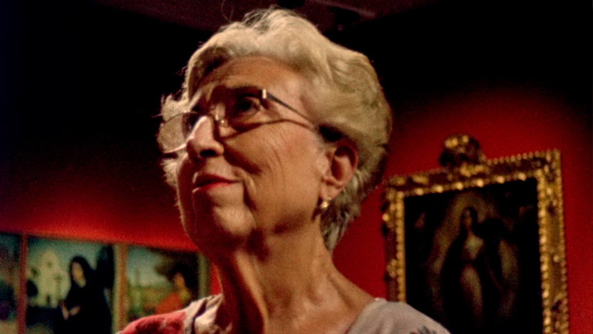 An elderly white lady with short white hair and glasses and red lipstick. Religious paintings in the background.