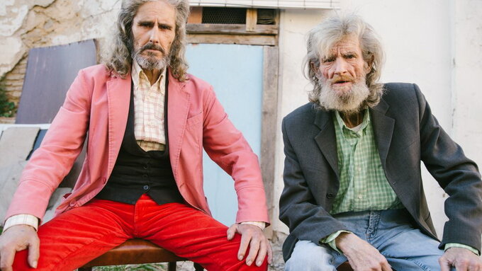 Two bearded white gentlemen with shoulder-length grey hair. They have their hands on their knees in a confident way.