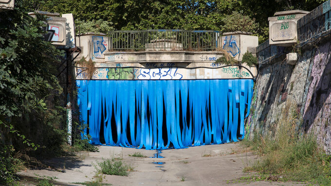 Bridge with graffiti. Tunnel below is covered with blue ribbons. Green trees in the background.