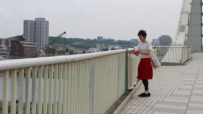 A woman in a red skirt and white blouse is standing on a bridge and holds a red thread that follows the bridge railing.