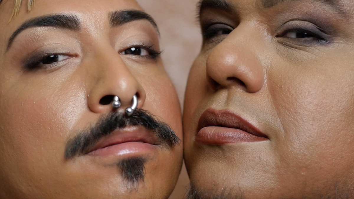 A close up of people looking directly into camera. One has a moustache and a nose piercing, one wears light makeup.