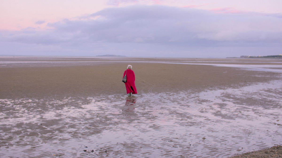 A woman in a red coat on a deserted beach. There is a wide expanse of sand with an island in the horizon.