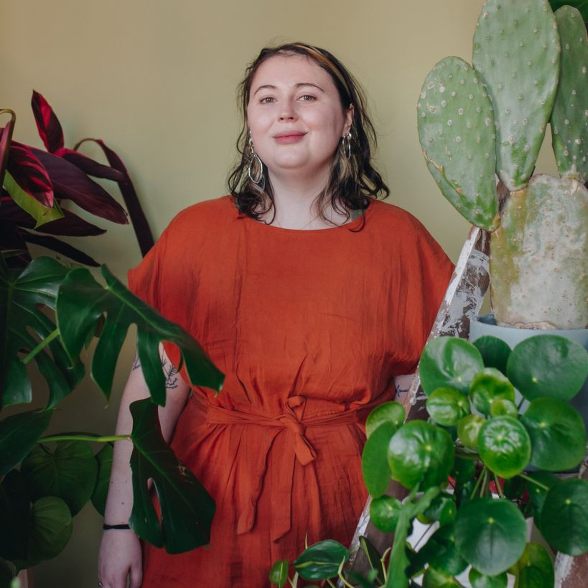 Emily May Armstrong, a person with dark hair wearing a red dress, standing within large plant foliage.