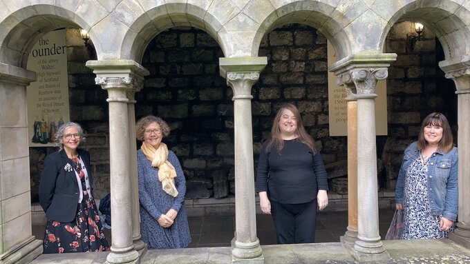 4 Women stand inside an old stone building. They each stand one by on under at an arched window.