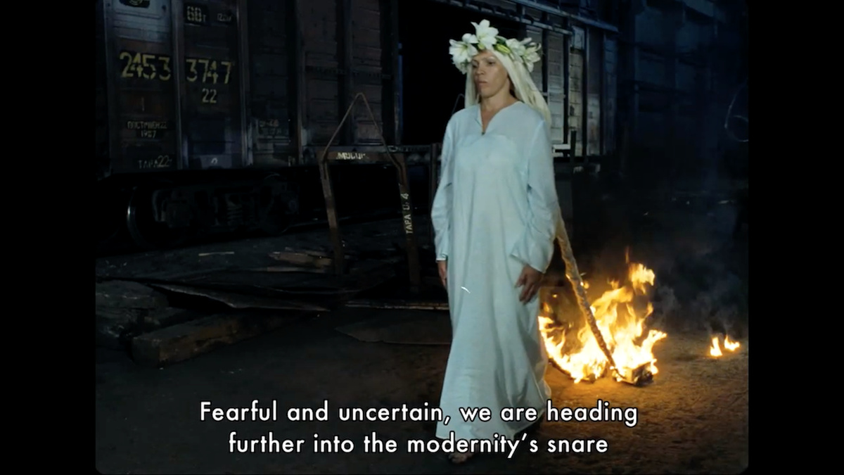 A woman in a long white dress with a headdress of flowers walks forward while her hair pulls two burning blocks of wood.