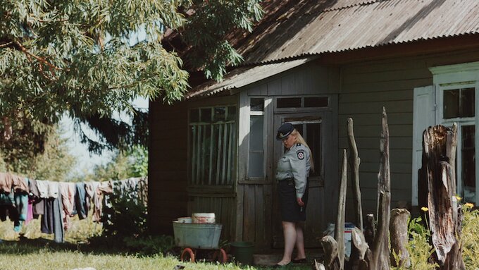 A police officer stands outside a rural country house bathed in sunshine.