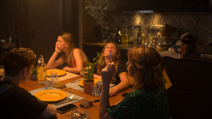 A mother and her teenage daughters sit around a dinner table smoking.
