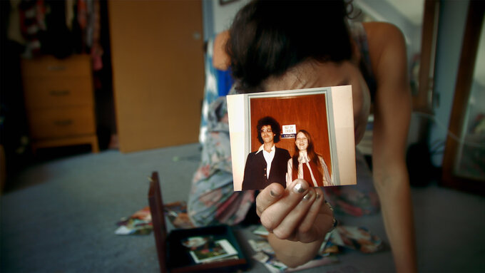 A person crouches on the floor and holds up an old photograph of a couple standing together.