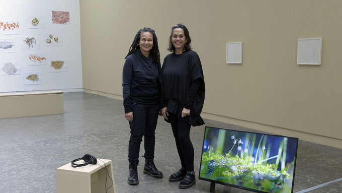 Annalee Davis and Amanda Thomson stand in the gallery space, they are both smiling, surrounded by their artwork.