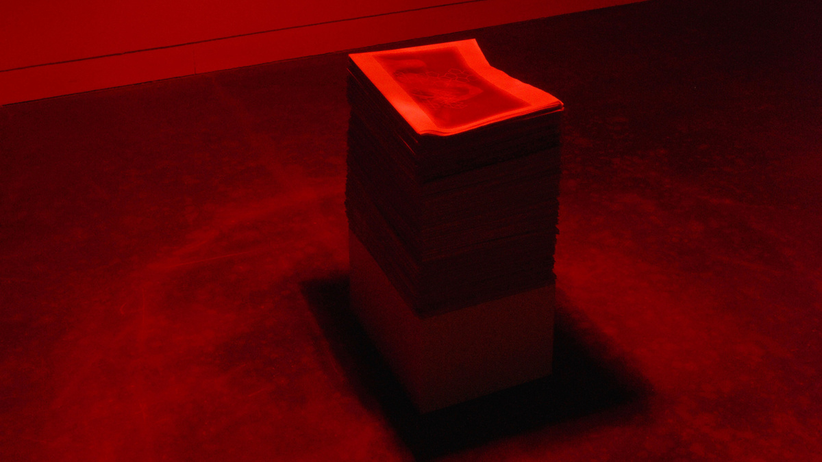 A stack of magazines lie on a gallery floor with a deep red light from overhead.