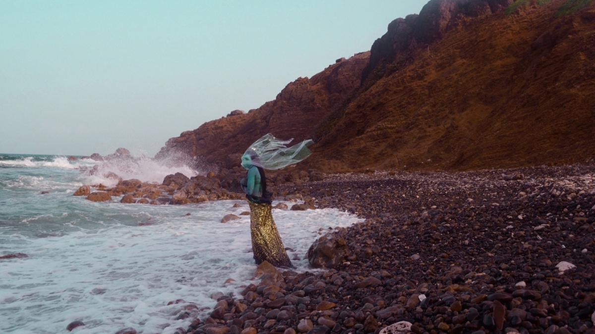 A lone figure stands on a rocky beach facing the sea, they wear a leopard print dress and clear veil.