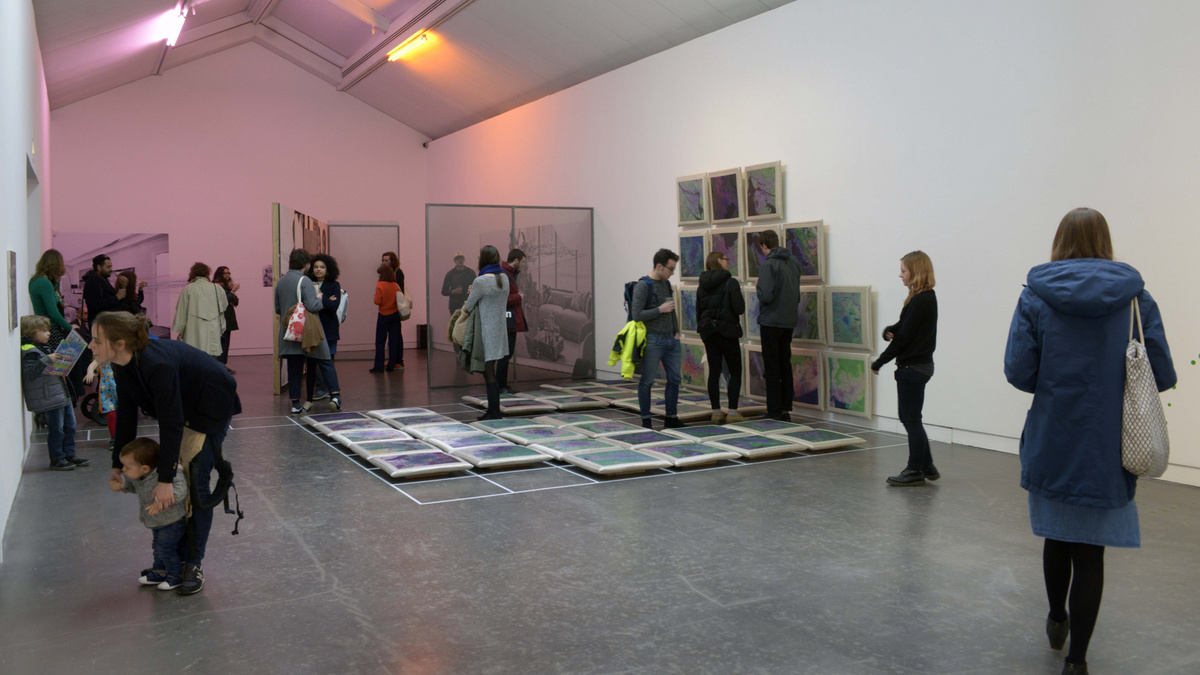 A group of people gather in a gallery space. Cushions are on the wall and floors, a purple light is at the far end.