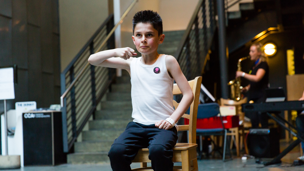 A photo of a young child mid-gesture on a chair in an open atrium space. Someone plays a saxophone in the background.