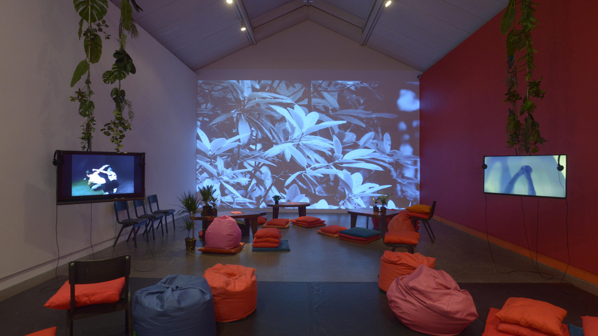 A photo of the interior of a gallery strewn with red beanbags, hanging foliage, video screens and a large projection.