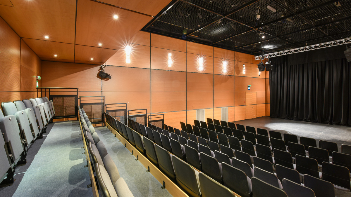 A photo of CCA well-lit theatre space from the top of the seating towards the stage. The walls are panelled in wood.