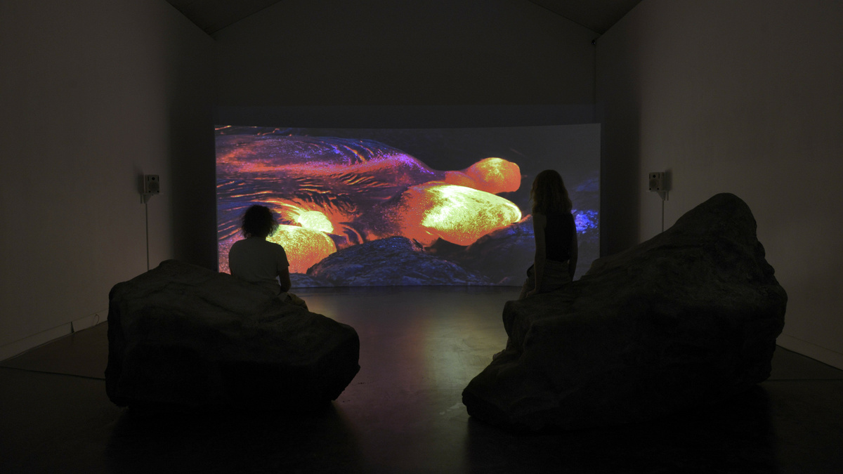 Two people sit on large rocks in a dark gallery space facing a large screen with a purple, yellow and red landscape.