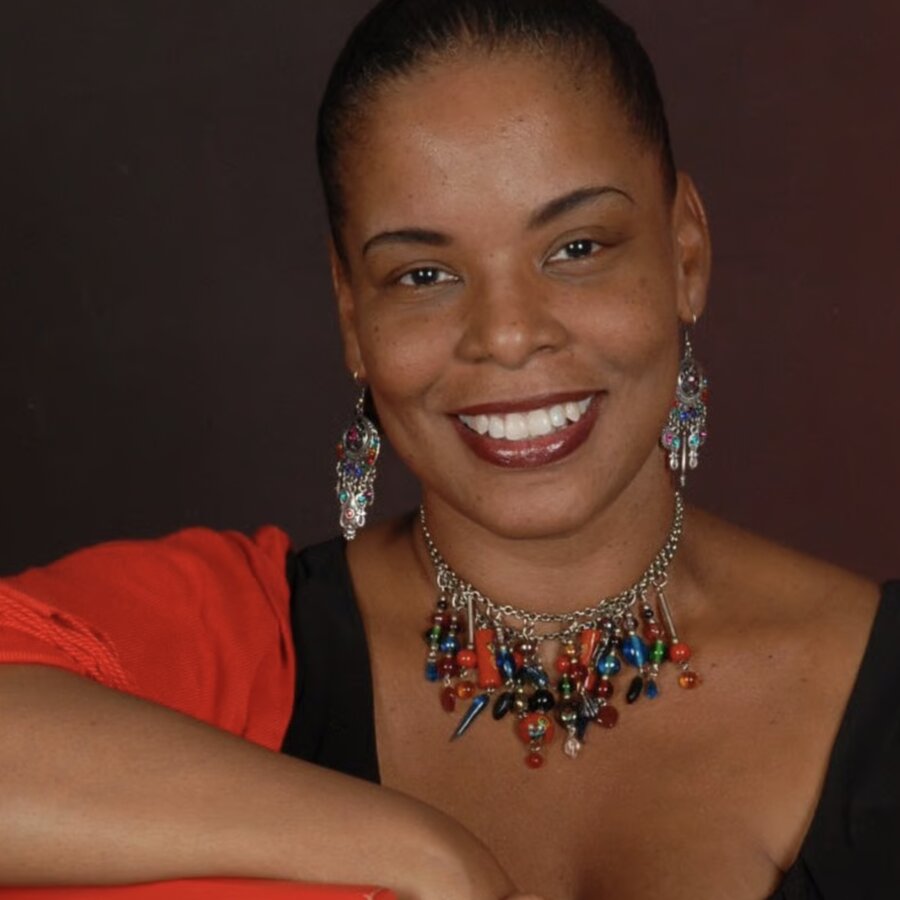 Muse is a black woman wearing a black dress and colourful jewellery. She is smiling at the camera in a welcoming pose.