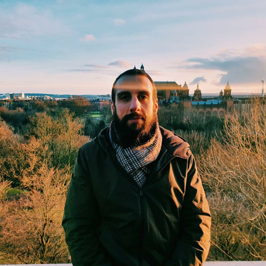 Faris Dannan wears a green jacket and patterned scarf. In the background are buildings and trees.