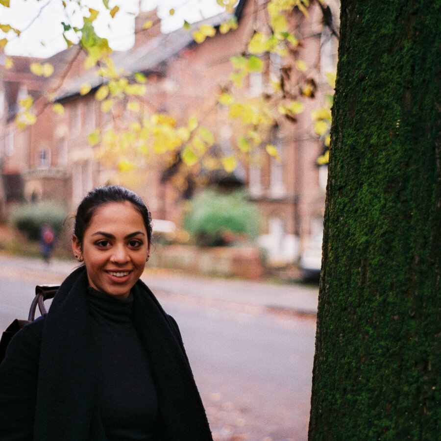 Aayushi Gupta wears a black shirt and jacket. In the background are red standstone tenements, trees and a white car.