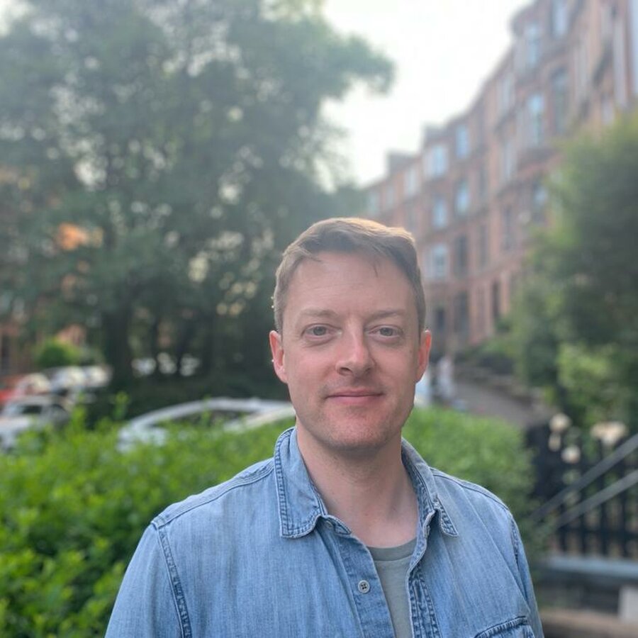 Martin Minton wears a blue shirt and grey t-shirt. In the background are sandstone terraced flats and trees.